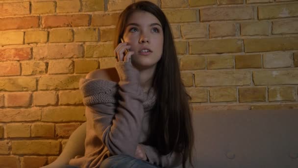 Portrait de jeune fille caucasienne assise sur le canapé et parlant sur smartphone souriant sur fond de maison confortable . — Video