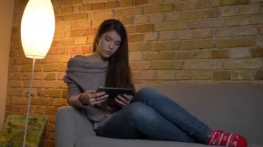 Tablet dikkatle izliyor ve rahat ev arka plan üzerinde bir helezon yapma kanepede oturan genç beyaz kız portresi.