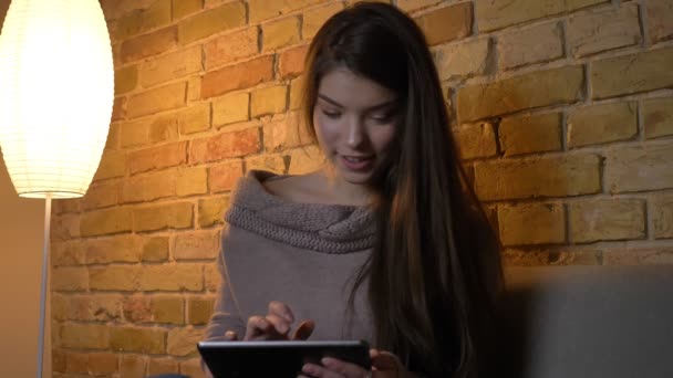 Portrait de jeune fille caucasienne assise sur le canapé regardant dans la tablette avec créativité et intérêt sur fond de maison confortable . — Video