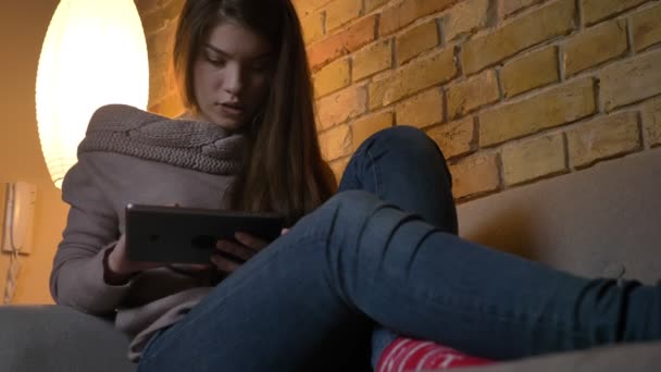Portrait from bottom of young caucasian girl sitting on sofa watching into tablet attentively on cosy home background. — Stock Video