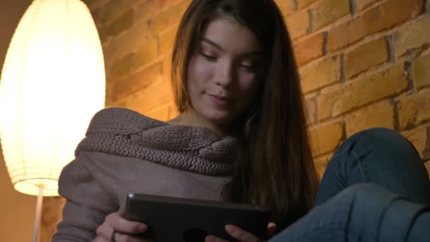 Portrait from bottom of young caucasian girl sitting on sofa watching into tablet thoughtfully on cosy home background. — Stock Video