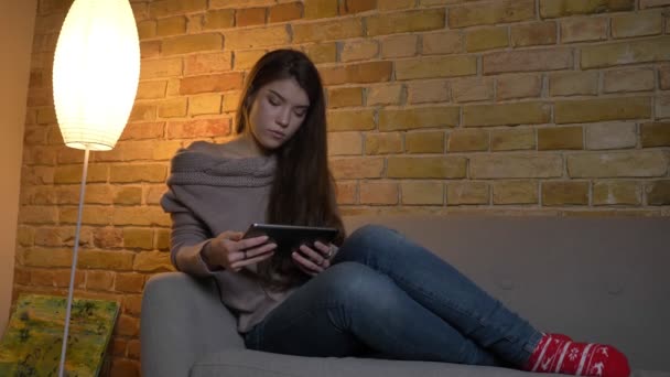 Retrato de una joven caucásica sentada en un sofá mirando a la tableta con atención y haciendo una coleta sobre un acogedor fondo casero . — Vídeo de stock