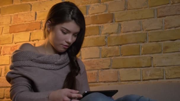 Portrait of young caucasian girl sitting on sofa watching into tablet attentively and making funny faces on cosy home background. — Stock Video