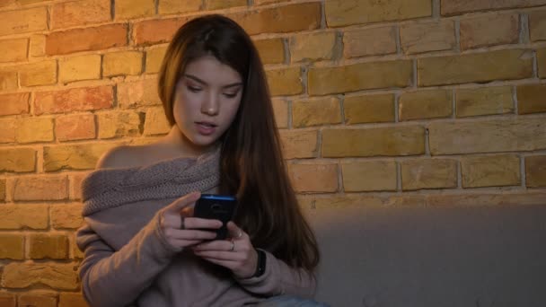 Portrait of young caucasian girl sitting on sofa and watching into the smartphone seriously on cosy home background. — Stock Video