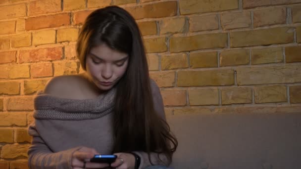 Portrait de jeune fille caucasienne concentrée assise sur le canapé et regardant sérieusement dans le smartphone sur fond de maison confortable . — Video