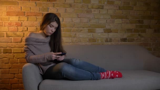 Concentré fille caucasienne assis sur le canapé et regarder dans le smartphone soigneusement sur fond de maison confortable . — Video