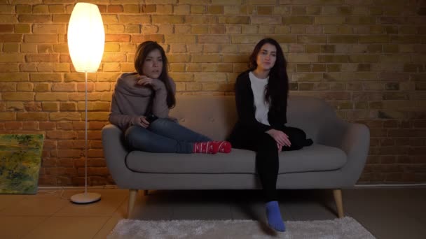 Retrato de chicas caucásicas jóvenes sentadas en el sofá y viendo la televisión con atención en el acogedor fondo de la casa . — Vídeos de Stock