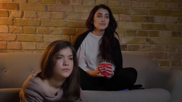 Retrato de cerca de dos jóvenes caucásicas sentadas en la sala de estar y viendo la televisión con diferentes emociones . — Vídeo de stock