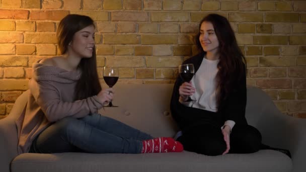 Retrato de jóvenes amigas caucásicas sentadas en un sofá con copas de vino y viendo películas en un acogedor ambiente hogareño . — Vídeo de stock