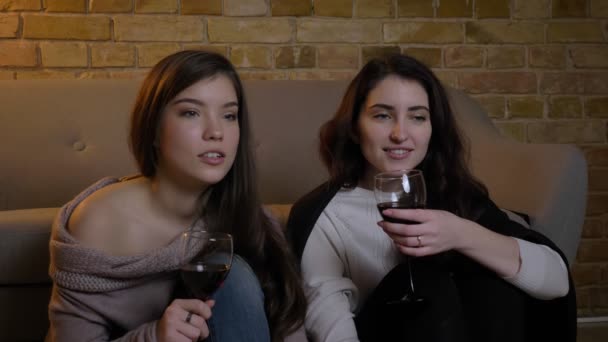 Portrait de jeunes amies caucasiennes assises par terre avec des verres de vin et regardant un film dans une atmosphère chaleureuse à la maison . — Video