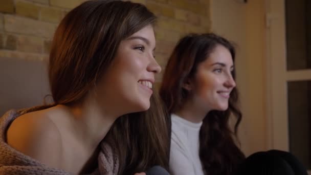 Ritratto ravvicinato di profilo di giovani ragazze caucasiche gioiose che guardano la TV e ridono su un accogliente sfondo domestico . — Video Stock