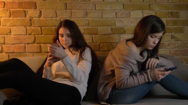 Νέοι Καυκάσιος φίλες κάθονται στον καναπέ και βλέποντας σε smartphones τους προσεκτικά στην ζεστή ατμόσφαιρα του σπιτιού. — Αρχείο Βίντεο