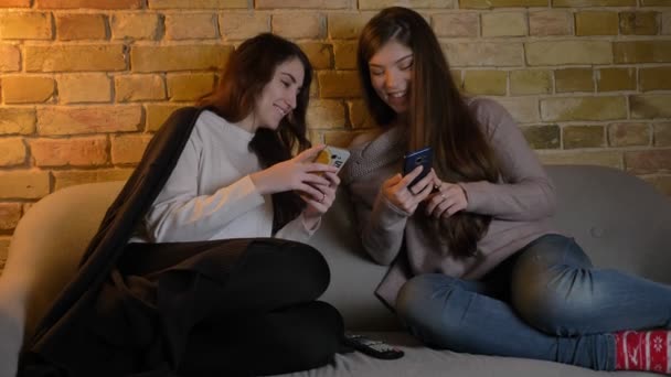 Retrato de jóvenes amigas caucásicas sentadas en el sofá y mirando en sus teléfonos inteligentes con alegría en un acogedor ambiente hogareño . — Vídeos de Stock