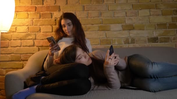 Retrato de chicas caucásicas jóvenes relajándose en el sofá y mirando en sus teléfonos inteligentes en un acogedor ambiente hogareño . — Vídeos de Stock
