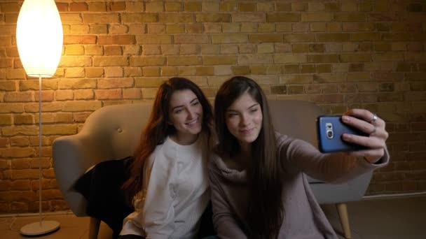 Portrait of young caucasian girls sitting on the floor and making joyful selfie-photos using smartphone in cosy home atmosphere. — Stock Video