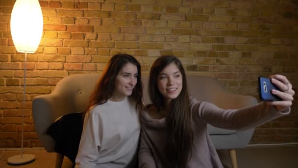 Retrato de jovens garotas caucasianas sentadas no chão e fazendo belas fotos de selfie usando smartphone em ambiente acolhedor em casa . — Vídeo de Stock