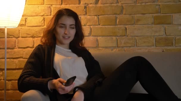 Portrait de jeune fille caucasienne avec les cheveux ondulés assis sur le canapé et regarder la télévision joyeusement sur fond de maison confortable . — Video