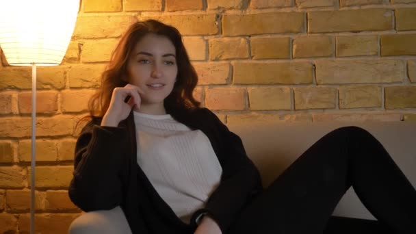 Portræt af ung kaukasisk pige med bølget hår sidder på sofaen og ser tv med stor entusiasme på hyggelig hjemmebaggrund . – Stock-video