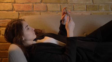 Koltukta yalan ve smartphone içinde rahat ev atmosferi içine izlerken dalgalı saçlı genç beyaz kız portresi.