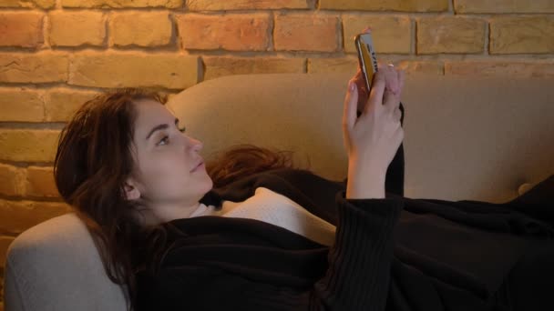Portret van de jonge Kaukasische meisje met golvende haren liggend op de bank kijken naar smartphone draait om de camera in huis gezellige — Stockvideo