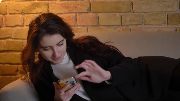 Брюнетка кавказская девушка с волнистыми волосами, лежащими на диване часы в смартфон и превращается в камеру в уютной домашней атмосфере . — стоковое видео