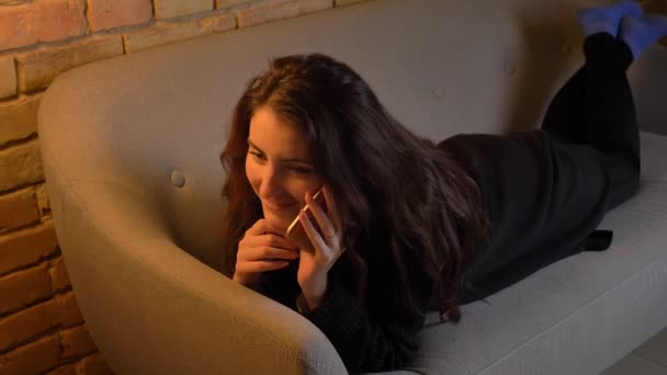 Ritratto di giovane ragazza caucasica con capelli ondulati sdraiata sul divano e che parla volentieri su smartphone in un'atmosfera accogliente a casa . — Video Stock