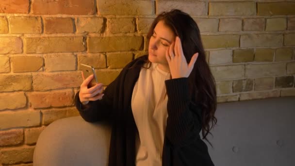 Молодая кавказская девушка с волнистыми волосами сидит на диване и делает селфи-фото улыбаясь на смартфоне в уютной домашней атмосфере . — стоковое видео