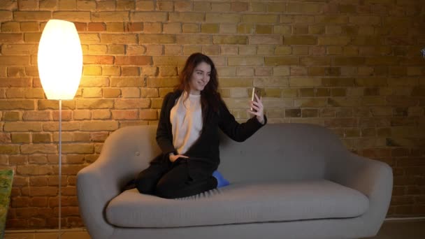 Joven chica caucásica con el pelo ondulado sentado en el sofá y haciendo selfie-foto felizmente en el teléfono inteligente en ambiente acogedor hogar . — Vídeos de Stock