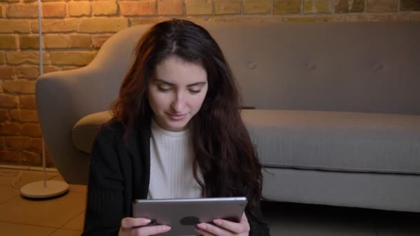 Retrato de una joven caucásica con el pelo ondulado sentado en el suelo observando atentamente en la tableta con alegría en un acogedor ambiente hogareño . — Vídeos de Stock