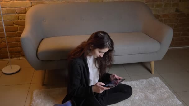 Chica caucásica con cabello ondulado morena sentada en el suelo mirando atentamente en la tableta en ambiente acogedor hogar . — Vídeos de Stock