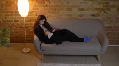 Koltukta yalan ve rahat ev ortamında tablet içine dikkatle izliyor dalgalı saçlı genç beyaz kız.