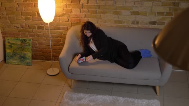 Joven chica caucásica con el pelo ondulado tumbado en el sofá y mirando atentamente en la tableta sale de la habitación . — Vídeos de Stock