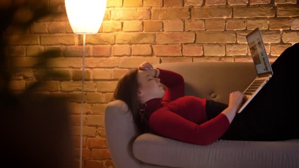 Nahaufnahme einer jungen attraktiven kaukasischen Frau, die sich ein Video auf dem Laptop ansieht, während sie drinnen auf der Couch liegt — Stockvideo