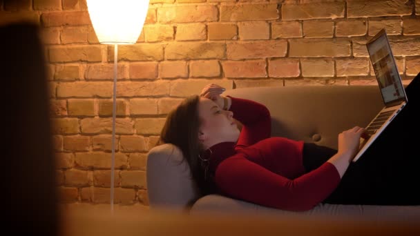 Крупним планом знімок молодої досить кавказької жінки, що дивиться фільм на ноутбуці, лежачи на дивані в приміщенні — стокове відео