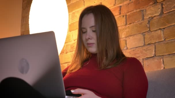 Nahaufnahme einer jungen hübschen kaukasischen Frau, die auf dem Laptop tippt, während sie drinnen auf dem Sofa sitzt — Stockvideo