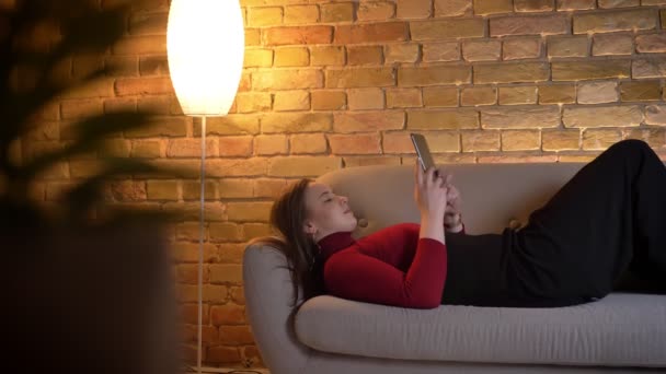 Nahaufnahme einer jungen hübschen kaukasischen Frau, die auf dem Tablet SMS schreibt, während sie drinnen auf der Couch liegt — Stockvideo