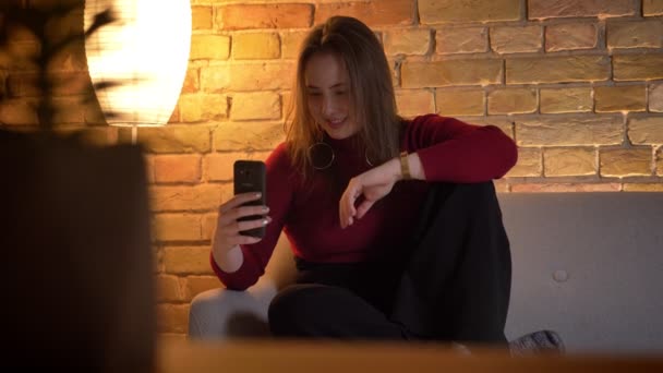 Крупним планом знімок молодої досить кавказької жінки, що має випадкову розмову через відеодзвінок по телефону, сидячи на дивані в приміщенні — стокове відео