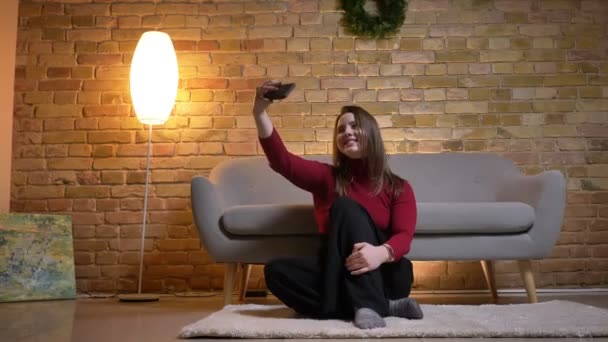 Closeup schieten van jonge mooie Kaukasische vrouw nemen van selfies op de telefoon en glimlachend zittend op de vloer poseren voor de camera — Stockvideo