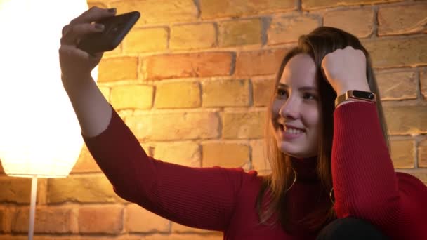 Vértes lő a fiatal szép kaukázusi nő, figyelembe véve a telefon mosolyogva selfies és hunyorítva beltéri — Stock videók