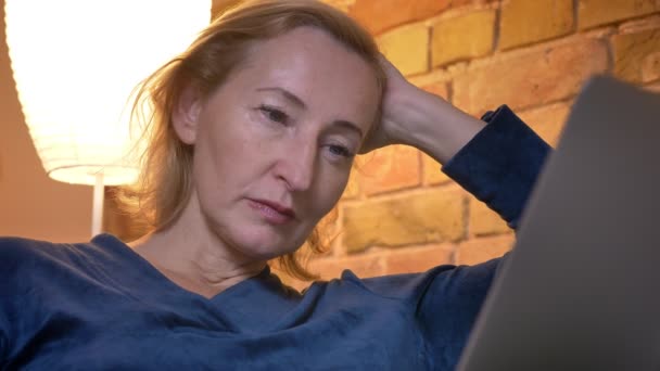 Крупним планом знімок літньої білої жінки, яка дивиться шоу на ноутбук уважно сидить на дивані в приміщенні — стокове відео