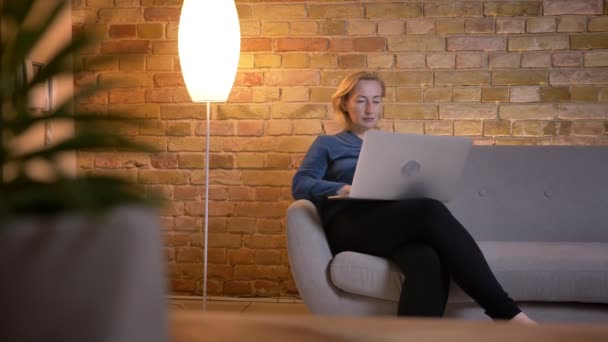Съемки крупным планом пожилой кавказки, печатающей на ноутбуке, сидящей на диване в помещении — стоковое видео