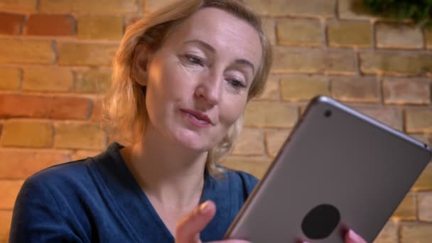 Nahaufnahme einer älteren kaukasischen Frau, die auf dem Tablet SMS schreibt, dann in die Kamera schaut und fröhlich lächelt — Stockvideo