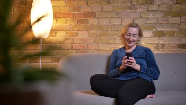Съемки крупным планом старшей кавказской женщины с помощью телефона, улыбающейся и смеющейся сидя на диване в помещении — стоковое видео