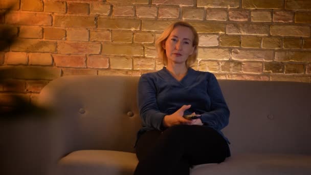 Κοντινό πλάνο της λευκής γυναίκας που βλέπει μια τηλεόραση να συρτάρει στον καναπέ σε ένα άνετο διαμέρισμα σε εσωτερικούς χώρους — Αρχείο Βίντεο