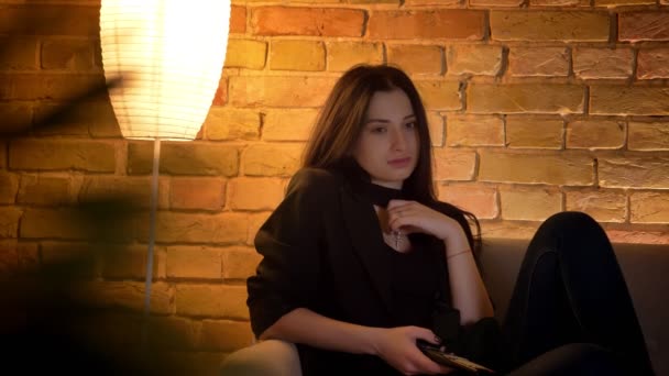 Młoda brunetka kaukaski dziewczyna siedzi na kanapie oglądania smutny film i płacz skromnie w przytulnej atmosferze domowej. — Wideo stockowe