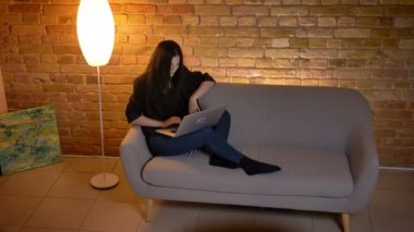 Genç esmer beyaz kız kanepede oturan ve ev atmosferi memnuniyetle laptop yazarak.