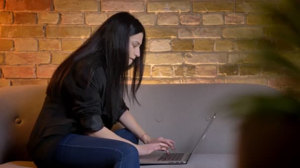 Junge brünette kaukasische Mädchen im Profil sitzt auf dem Sofa und tippt auf dem Laptop aufmerksam und gerne in häuslicher Atmosphäre. — Stockvideo