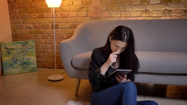 Retrato de la joven morena caucásica sentada en el suelo y trabajando con la tableta con atención en el ambiente del hogar . — Vídeos de Stock