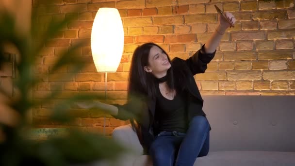 Ritratto di giovane ragazza caucasica bruna seduta sul divano e che parla in videochat utilizzando lo smartphone in atmosfera domestica . — Video Stock