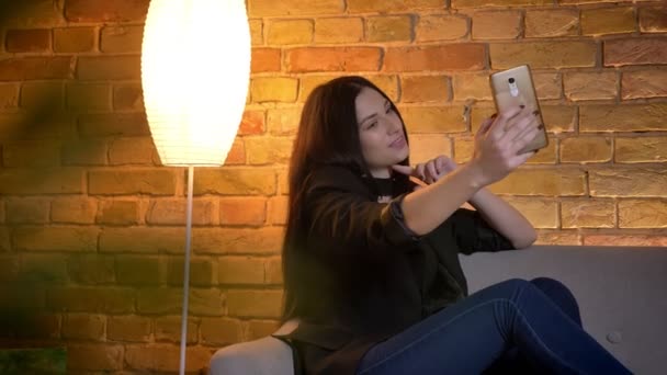 Портрет юной брюнетки кавказской девушки, сидящей на диване и делающей селфи-фотографии в домашней обстановке . — стоковое видео